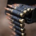 The image shows a close-up photograph of a linked ammunition belt, commonly used with machine guns or automatic weapons. The cartridges are held together by dark metal links in a standard disintegrating belt configuration. The copper-tipped projectiles and brass casings are clearly visible in sharp detail.