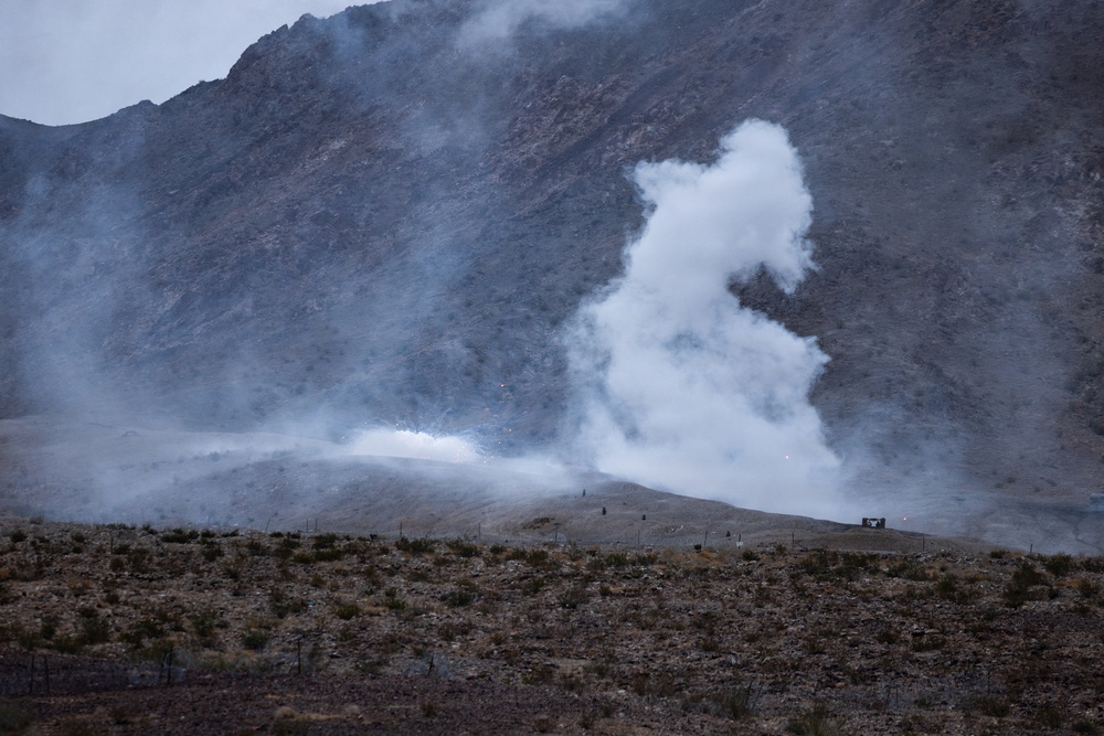 The image shows a large plume of white smoke rising from a hillside in an arid, mountainous landscape. The smoke appears to originate from a specific point on a ridge, suggesting a controlled explosion, detonation, or ordnance disposal activity. Small figures and what appears to be a structure are visible near the base of the smoke column, indicating human presence at the site.