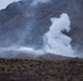 The image shows a large plume of white smoke rising from a hillside in an arid, mountainous landscape. The smoke appears to originate from a specific point on a ridge, suggesting a controlled explosion, detonation, or ordnance disposal activity. Small figures and what appears to be a structure are visible near the base of the smoke column, indicating human presence at the site.