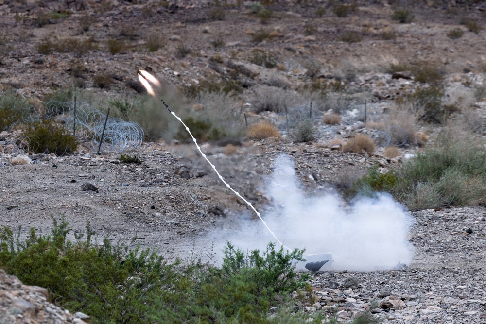 A rocket or missile has been launched from ground level in an arid, desert-like terrain. A white smoke trail connects the launch point to the projectile, which is visible in the upper left with a bright flame. The surrounding landscape features sparse desert vegetation, rocky ground, and what appears to be concertina wire fencing.