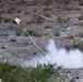 A rocket or missile has been launched from ground level in an arid, desert-like terrain. A white smoke trail connects the launch point to the projectile, which is visible in the upper left with a bright flame. The surrounding landscape features sparse desert vegetation, rocky ground, and what appears to be concertina wire fencing.