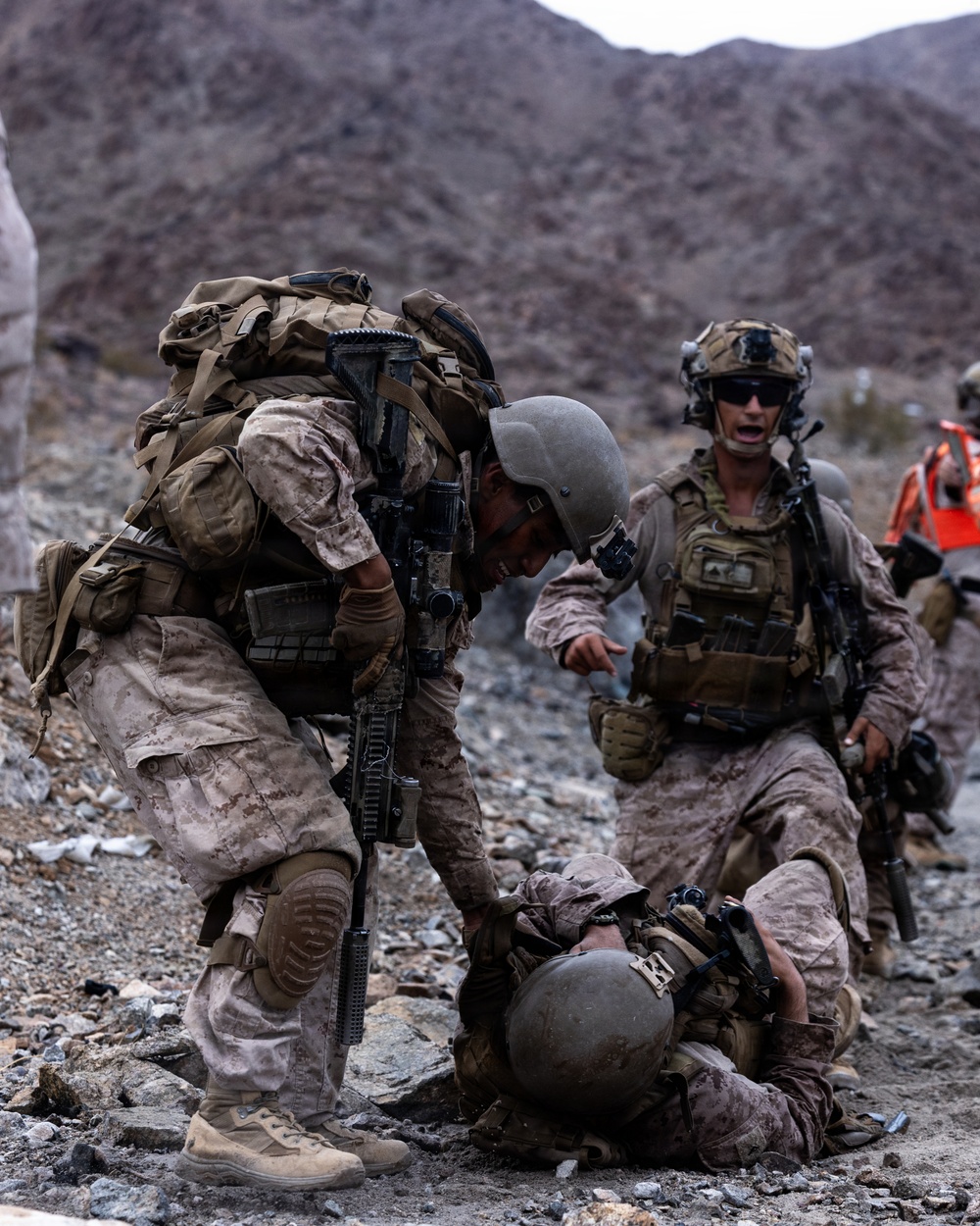 The image depicts what appears to be U.S. Marines in desert MARPAT camouflage conducting a casualty assistance or combat training exercise in a rocky, arid mountainous terrain. One fully-equipped Marine is bending down to assist a downed Marine on the ground, while a second standing Marine in tactical gear appears to be issuing commands or coordinating the response. The presence of an orange marker or safety flag in the background suggests this may be a training exercise rather than a live combat scenario.