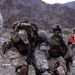 The image depicts what appears to be U.S. Marines in desert MARPAT camouflage conducting a casualty assistance or combat training exercise in a rocky, arid mountainous terrain. One fully-equipped Marine is bending down to assist a downed Marine on the ground, while a second standing Marine in tactical gear appears to be issuing commands or coordinating the response. The presence of an orange marker or safety flag in the background suggests this may be a training exercise rather than a live combat scenario.