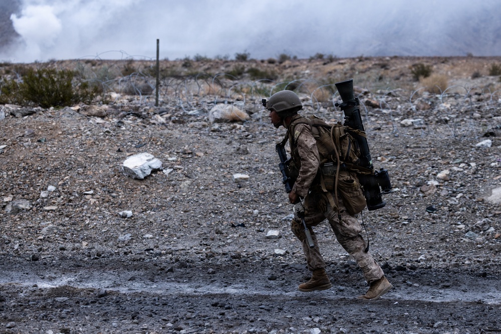 A single soldier in desert camouflage uniform is walking across rocky, arid terrain carrying a large weapon system on their back, consistent with a man-portable anti-tank or rocket launcher. The background shows a barbed wire perimeter, rocky desert landscape, and smoke or dust clouds rising in the distance. The scene appears to be an active military training exercise or combat operation.