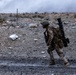 A single soldier in desert camouflage uniform is walking across rocky, arid terrain carrying a large weapon system on their back, consistent with a man-portable anti-tank or rocket launcher. The background shows a barbed wire perimeter, rocky desert landscape, and smoke or dust clouds rising in the distance. The scene appears to be an active military training exercise or combat operation.
