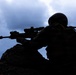 A silhouetted soldier in full tactical gear is positioned behind a rocky outcrop, aiming a scoped rifle with a suppressor attached. The image is a dramatic backlit shot against an overcast, cloudy sky, rendering the subject almost entirely in dark silhouette. The composition emphasizes a defensive or overwatch firing position.