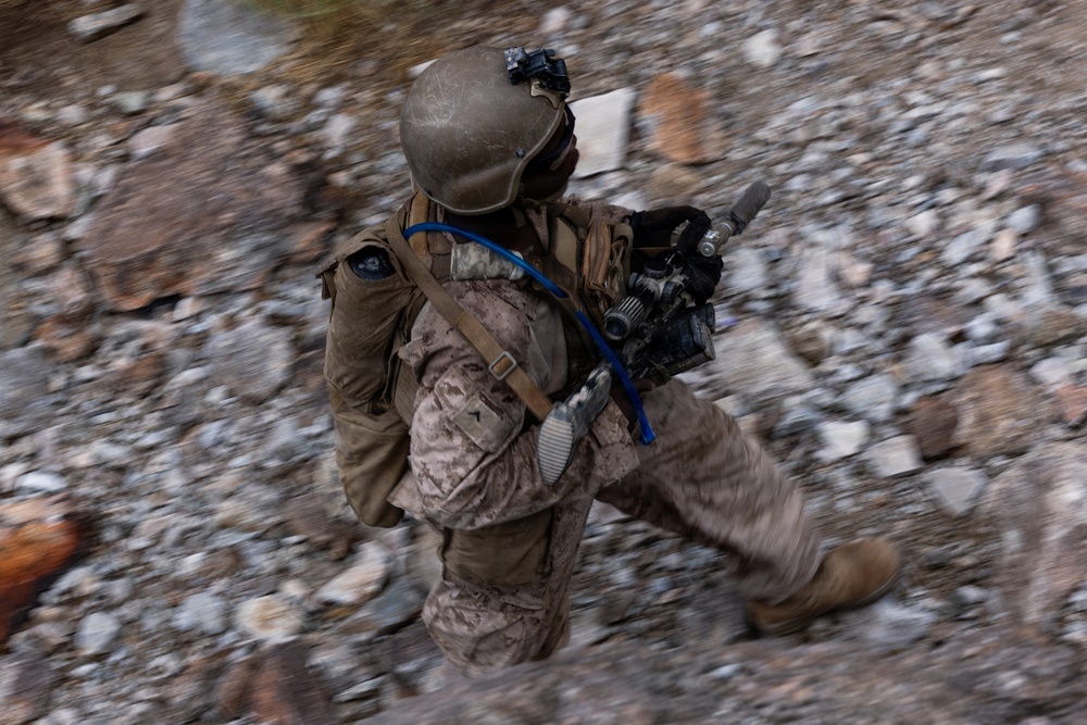 A military service member in desert camouflage (MARPAT pattern) is captured mid-movement across rocky terrain, photographed from an elevated angle. The soldier is fully equipped with tactical gear, a helmet with mounted accessories, and is carrying a weapon with optics. The image uses motion blur to convey speed and urgency of movement.