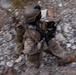 A military service member in desert camouflage (MARPAT pattern) is captured mid-movement across rocky terrain, photographed from an elevated angle. The soldier is fully equipped with tactical gear, a helmet with mounted accessories, and is carrying a weapon with optics. The image uses motion blur to convey speed and urgency of movement.