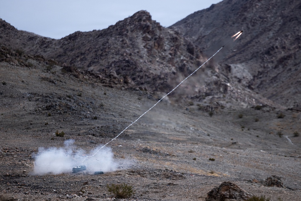 A ground-launched projectile or rocket is captured mid-flight in a desert mountain environment, leaving a visible smoke trail from the launch point to the projectile in the upper right of the frame. White smoke is billowing from a small launch device on the rocky ground at lower left. The setting appears to be an arid, mountainous terrain consistent with a military test or training range.