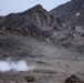A ground-launched projectile or rocket is captured mid-flight in a desert mountain environment, leaving a visible smoke trail from the launch point to the projectile in the upper right of the frame. White smoke is billowing from a small launch device on the rocky ground at lower left. The setting appears to be an arid, mountainous terrain consistent with a military test or training range.