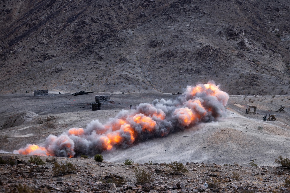 A large linear explosion or controlled detonation is occurring across a desert/arid mountainous terrain. Multiple fireballs and smoke clouds are visible in a sequential line pattern, suggesting a deliberate demolition or military exercise. The setting appears to be a rocky, barren hillside with some structures visible in the background.
