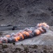 A large linear explosion or controlled detonation is occurring across a desert/arid mountainous terrain. Multiple fireballs and smoke clouds are visible in a sequential line pattern, suggesting a deliberate demolition or military exercise. The setting appears to be a rocky, barren hillside with some structures visible in the background.