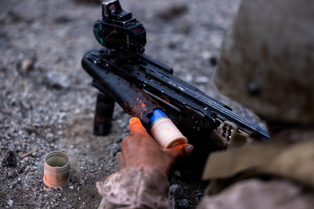 A military or law enforcement personnel is loading or reloading a grenade launcher, likely an underbarrel or standalone type, with what appears to be a practice/training round colored in orange and blue. The weapon is equipped with a red dot optic sight on top. Orange paint/marking powder residue is visible on the weapon and the shooter's hand, along with a spent canister on the ground, indicating a training exercise.