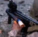 A military or law enforcement personnel is loading or reloading a grenade launcher, likely an underbarrel or standalone type, with what appears to be a practice/training round colored in orange and blue. The weapon is equipped with a red dot optic sight on top. Orange paint/marking powder residue is visible on the weapon and the shooter's hand, along with a spent canister on the ground, indicating a training exercise.