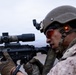 A U.S. Marine in full combat gear is aiming a rifle equipped with a magnified optic scope. The Marine is wearing MARPAT camouflage uniform, a PASGT-style helmet with a mounted device, and tactical gloves. The image is a close-up profile shot taken in an overcast outdoor environment.