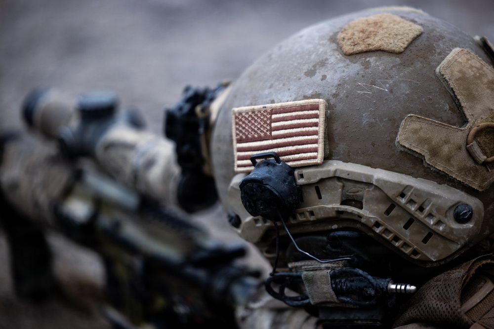 Close-up photograph of a US military soldier's helmet in the foreground, featuring a subdued American flag patch on the side. The soldier appears to be in a tactical position with a scoped rifle visible in the background alongside at least one other soldier. The image uses shallow depth of field, keeping the helmet in sharp focus while blurring the background elements.