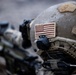 Close-up photograph of a US military soldier's helmet in the foreground, featuring a subdued American flag patch on the side. The soldier appears to be in a tactical position with a scoped rifle visible in the background alongside at least one other soldier. The image uses shallow depth of field, keeping the helmet in sharp focus while blurring the background elements.