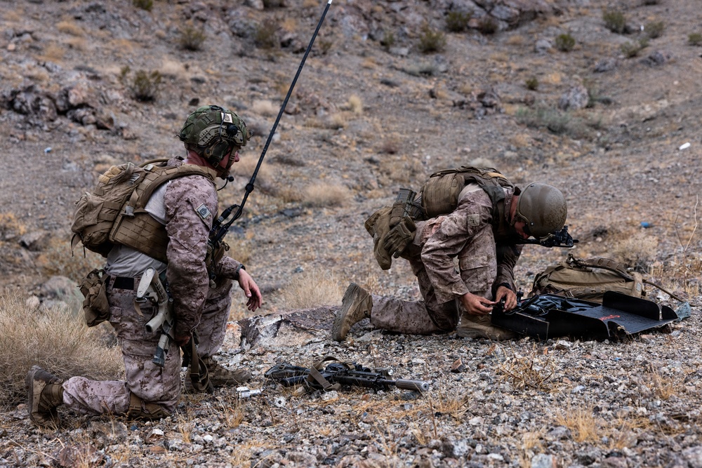 Two military personnel in desert camouflage uniforms are kneeling on rocky, arid terrain. The left individual appears to be communicating via radio with a tall antenna visible, while the right individual is operating or inspecting a piece of equipment. Both are fully equipped with tactical gear, body armor, and helmets.