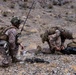 Two military personnel in desert camouflage uniforms are kneeling on rocky, arid terrain. The left individual appears to be communicating via radio with a tall antenna visible, while the right individual is operating or inspecting a piece of equipment. Both are fully equipped with tactical gear, body armor, and helmets.