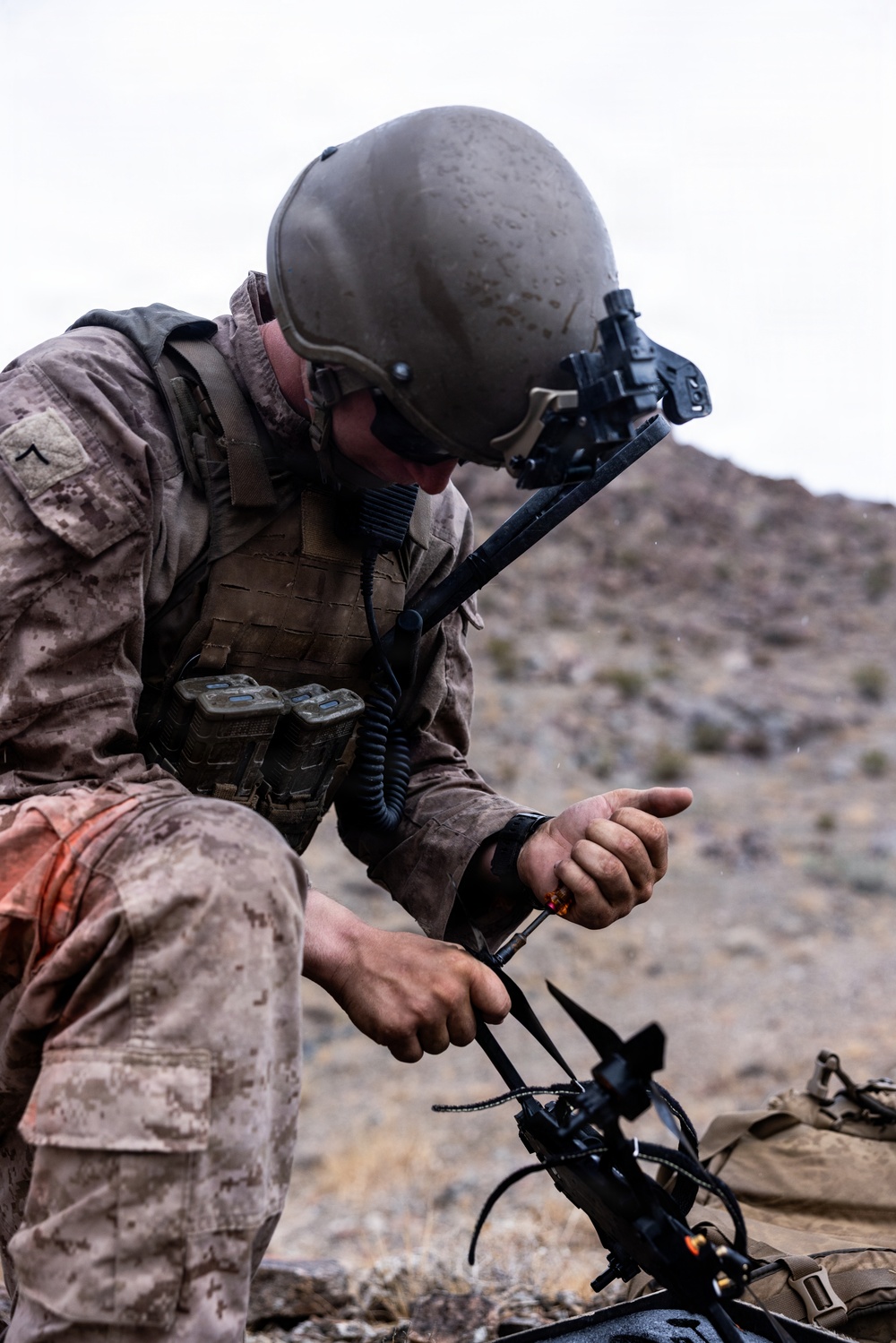 A military service member in desert camouflage uniform is crouched in an arid, rocky terrain working with communications or tactical equipment. The individual is wearing a combat helmet with a night vision device mount and a tactical vest with MOLLE webbing. The setting appears to be a desert training environment based on the landscape and equipment condition.