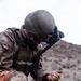 A military service member in desert camouflage uniform is crouched in an arid, rocky terrain working with communications or tactical equipment. The individual is wearing a combat helmet with a night vision device mount and a tactical vest with MOLLE webbing. The setting appears to be a desert training environment based on the landscape and equipment condition.