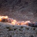 The image shows a series of controlled explosions occurring in a rocky, arid desert valley or canyon setting. Multiple simultaneous detonations are visible along what appears to be a linear path or road, producing large orange fireballs and dark gray smoke clouds. The setting and pattern of explosions suggest a military demolition exercise or mine-clearing operation.