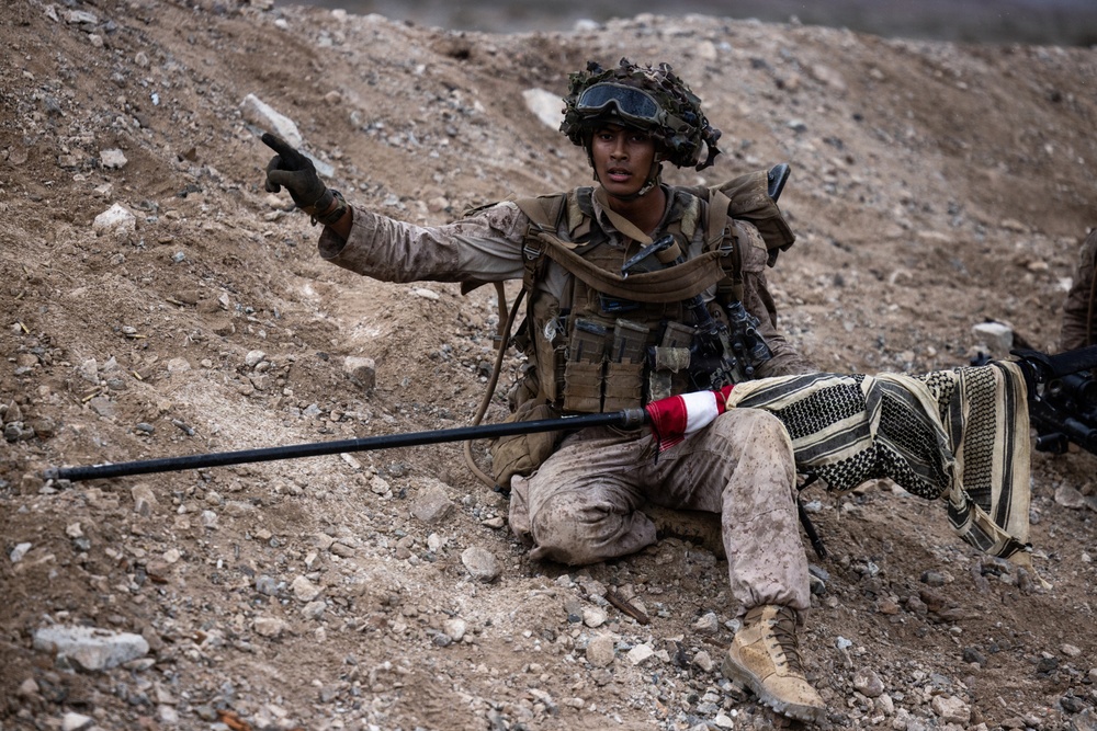 A soldier in full combat gear is seated on rocky, arid terrain while pointing in a direction, likely issuing tactical instructions or identifying a target. The individual is equipped with a sniper or precision rifle wrapped in a camouflage covering, tactical vest with magazine pouches, helmet with foliage camouflage, and protective goggles. The setting appears to be a desert or semi-arid training or operational environment.