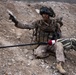 A soldier in full combat gear is seated on rocky, arid terrain while pointing in a direction, likely issuing tactical instructions or identifying a target. The individual is equipped with a sniper or precision rifle wrapped in a camouflage covering, tactical vest with magazine pouches, helmet with foliage camouflage, and protective goggles. The setting appears to be a desert or semi-arid training or operational environment.
