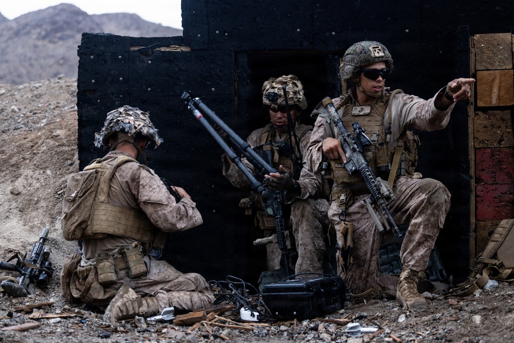 Three military personnel in desert camouflage uniforms are positioned against a damaged structure in an arid, rocky environment. One soldier is kneeling and appears to be working with equipment on the ground, while a second handles what appears to be a tripod-mounted device, and a third is pointing in a direction while holding a rifle. The scene appears to be a military training exercise or tactical operation.