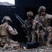 Three military personnel in desert camouflage uniforms are positioned against a damaged structure in an arid, rocky environment. One soldier is kneeling and appears to be working with equipment on the ground, while a second handles what appears to be a tripod-mounted device, and a third is pointing in a direction while holding a rifle. The scene appears to be a military training exercise or tactical operation.