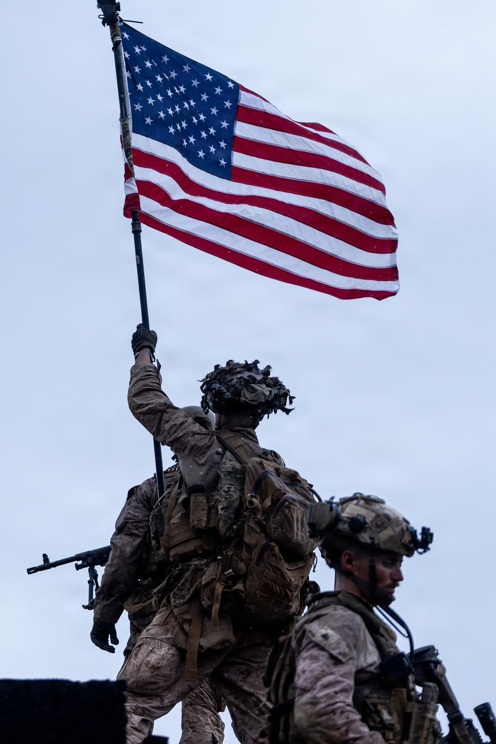 Two heavily equipped military personnel are photographed from below against an overcast sky. The soldier in the background is raising a large American flag on a pole with one arm extended overhead. Both individuals are wearing full combat gear including helmets, tactical vests, and carrying weapons.
