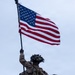 Two heavily equipped military personnel are photographed from below against an overcast sky. The soldier in the background is raising a large American flag on a pole with one arm extended overhead. Both individuals are wearing full combat gear including helmets, tactical vests, and carrying weapons.
