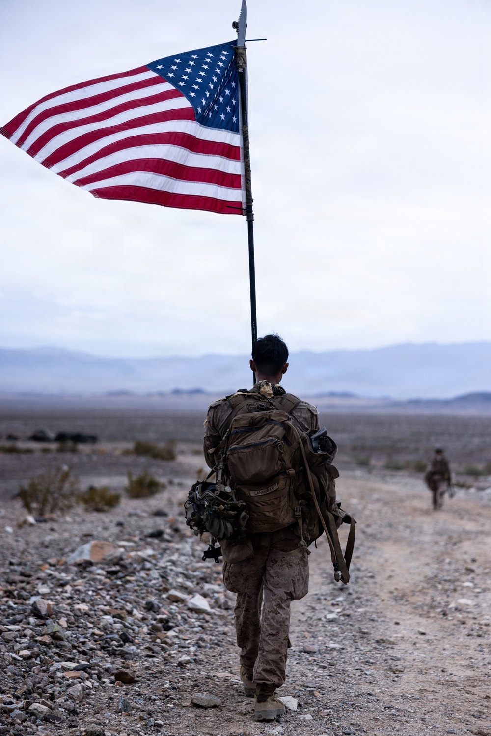 A U.S. Marine in desert MARPAT camouflage carries a large American flag on a pole while conducting a ruck march across an arid, rocky desert terrain. The soldier bears a heavy tactical backpack and is moving along a dirt trail with mountains visible in the background. A second service member is visible in the distance ahead on the same trail.
