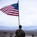 A U.S. Marine in desert MARPAT camouflage carries a large American flag on a pole while conducting a ruck march across an arid, rocky desert terrain. The soldier bears a heavy tactical backpack and is moving along a dirt trail with mountains visible in the background. A second service member is visible in the distance ahead on the same trail.