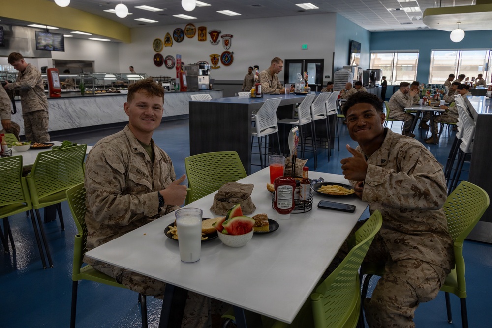 Two U.S. Marines in MARPAT camouflage uniforms are seated at a dining table in a military mess hall (chow hall), giving thumbs-up gestures toward the camera. Their table holds various food items including watermelon, french fries, a burger, milk, and a red beverage. The background shows a modern, well-lit dining facility with multiple other service members eating and a food service counter with military unit insignia patches displayed on the wall.
