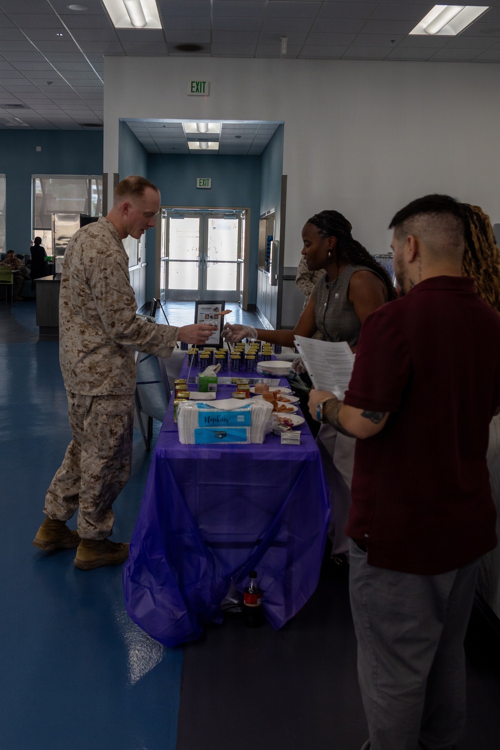 A food sampling or information booth event is taking place inside what appears to be a military dining facility or cafeteria. A person in U.S. Marine Corps desert camouflage uniform is interacting with two civilian staff members behind a purple-tablecloth-covered table. The table is set up with various food samples, napkins, small containers, and informational materials.