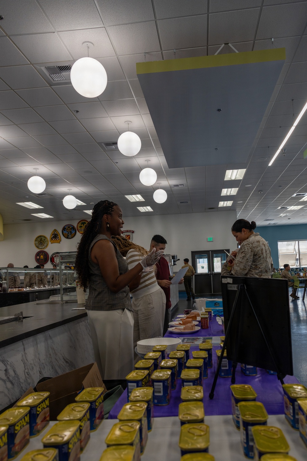 The image shows an indoor food service or tasting event, likely at a military dining facility, with numerous cans of SPAM arranged on a purple-covered table in the foreground. Several individuals, including one wearing disposable gloves, are engaged in what appears to be a food demonstration or sampling event. Military unit insignia patches are visible on the wall in the background, suggesting a military mess hall or chow hall setting.