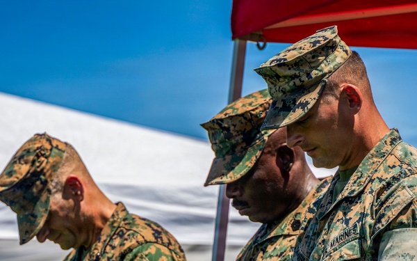 A group of U.S. Marines in MARPAT woodland camouflage uniforms stand with heads bowed, likely during a prayer or moment of silence at an outdoor ceremony. The setting appears to be a formal military event held under tents on a sunny day. Multiple service members are visible, with name tapes reading 'MARINES' on their uniforms.