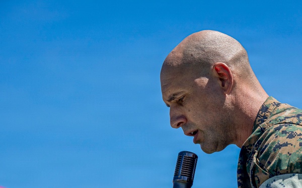 A bald male individual wearing United States Marine Corps MARPAT (woodland) camouflage uniform is speaking into a handheld microphone outdoors. He appears to be addressing an audience at a formal or ceremonial event, leaning forward with intensity. A red canopy tent is partially visible in the lower left foreground against a clear blue sky.