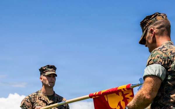 Two U.S. Marines in MARPAT digital camouflage uniforms are participating in a formal military ceremony involving the transfer of a unit guidon (flag). The red and yellow flag appears to bear Marine Corps insignia and unit designation markings. The ceremony takes place outdoors with a formation of Marines visible in the background, set against a mountainous tropical landscape consistent with Hawaii.