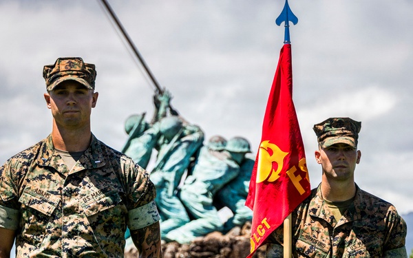 A formal U.S. Marine Corps military ceremony is taking place outdoors, with Marines in MARPAT camouflage uniforms standing at attention. Two Marines are prominently positioned in the foreground, one holding a red unit guidon flag. Behind them stands a replica of the iconic Iwo Jima Memorial statue, suggesting this event is taking place at a Marine Corps installation, likely in Hawaii based on the visible mountain terrain.