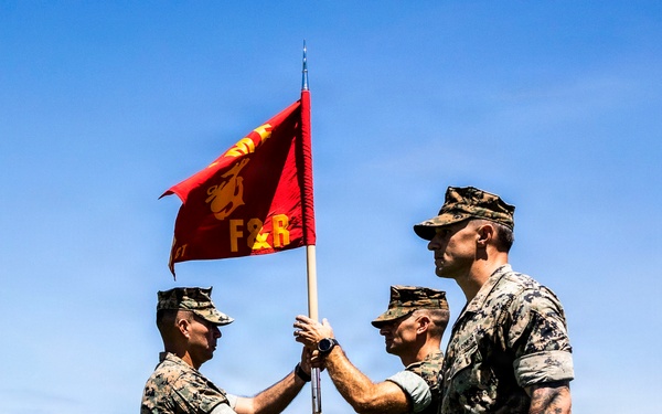 Three U.S. Marines in MARPAT camouflage uniforms are conducting a guidon transfer ceremony on a grass field. Two Marines are exchanging a red guidon flag bearing the letters 'F&amp;R' and a Marine Corps eagle, globe, and anchor emblem. A static display aircraft marked 'MARINES' is visible in the background, suggesting a Marine Corps installation.