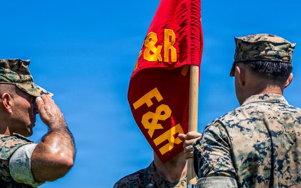 Three military personnel in U.S. Marine Corps MARPAT camouflage uniforms are participating in what appears to be a formal military ceremony. One Marine on the left is rendering a salute, while a central figure holds a red guidon flag bearing the letters 'F&amp;R' in yellow. A third Marine stands with his back to the camera on the right, displaying a visible tattoo on his forearm.