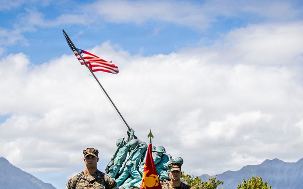 A formal U.S. Marine Corps military ceremony is taking place outdoors in front of a replica of the Iwo Jima Memorial statue, with an American flag raised above it. Numerous Marines in camouflage utilities are assembled in formation, with two prominent figures standing at the front — one holding a red Marine Corps unit guidon. The setting appears to be a tropical military installation, likely in Hawaii, based on the distinctive mountain ridgeline in the background.