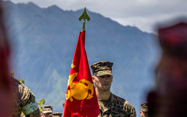 A U.S. Marine in woodland MARPAT camouflage uniform stands at attention holding a red guidon flag marked '3rd LCT' (Landing Craft Team) with the Marine Corps Eagle, Globe, and Anchor emblem. The scene appears to be a formal military ceremony or formation, with multiple Marines visible in the background. The setting features a dramatic mountainous backdrop, consistent with locations such as Hawaii (Kaneohe Bay/MCB Hawaii).