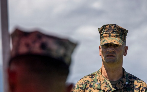 A U.S. Marine officer, identified by name tape as 'Wallace,' stands in the center of the frame wearing MARPAT woodland digital camouflage uniform and cover. He appears to be addressing or observing a group of Marines, with two out-of-focus Marines visible in the foreground. The setting appears to be an outdoor location with mountains and cloudy skies visible in the background.