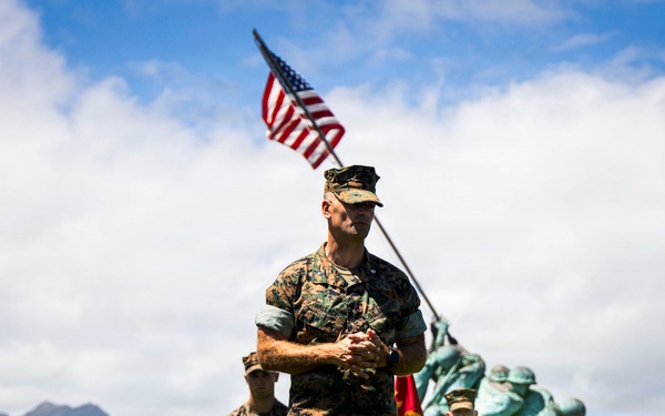 A group of U.S. Marines in woodland MARPAT camouflage uniforms are assembled on a grassy field with mountainous terrain in the background, consistent with a Hawaiian landscape. A prominent Marine in the foreground stands with arms clasped while an American flag flies behind him. In the mid-ground, a teal/patina-colored statue resembling the Iwo Jima Memorial flag-raising is visible, along with a red Marine Corps guidon.