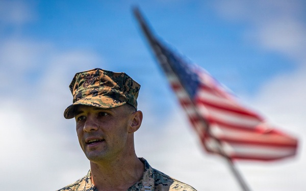 A U.S. Marine in woodland MARPAT digital camouflage uniform stands in the foreground with an American flag visible and slightly out of focus in the background. The Marine's name tape reads 'FITZPATRICK' and the branch tape reads 'U.S. MARINES'. The image appears to be a professional or official military photograph taken outdoors under a partly cloudy sky.