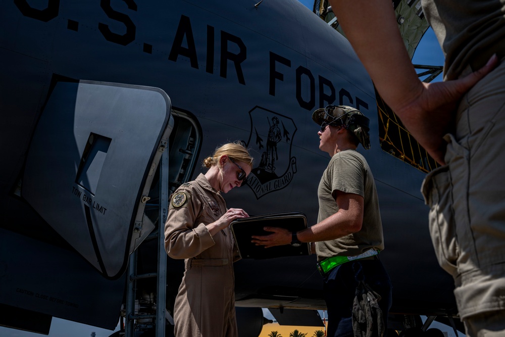 Two U.S. Air Force personnel are conducting a pre-flight or post-flight inspection beside a large military aircraft marked 'U.S. AIR FORCE' with a National Guard insignia. A woman in a flight suit is reviewing documents on a clipboard held by a second person in a t-shirt and cap. The scene appears to take place on a flight line in a sunny, arid environment.