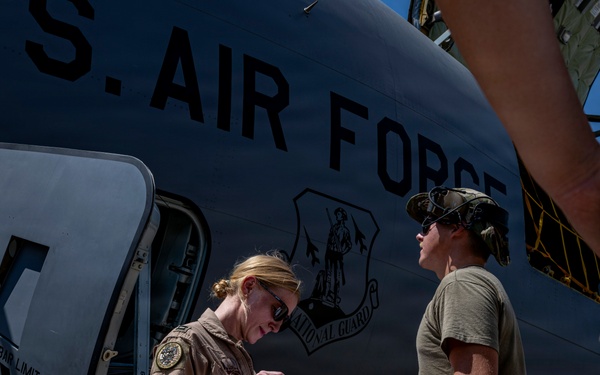 Two U.S. Air Force personnel are conducting a pre-flight or post-flight inspection beside a large military aircraft marked 'U.S. AIR FORCE' with a National Guard insignia. A woman in a flight suit is reviewing documents on a clipboard held by a second person in a t-shirt and cap. The scene appears to take place on a flight line in a sunny, arid environment.